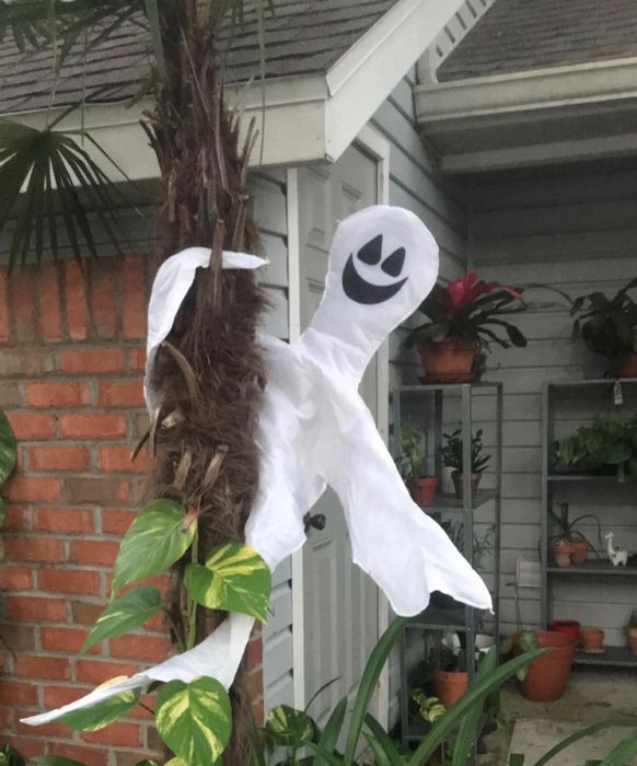 A cheerful ghost decoration hangs from a tree in front of a house, surrounded by lush greenery