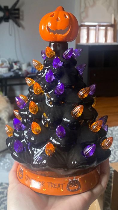 A ceramic Halloween tree adorned with orange and purple lights, featuring a pumpkin topper and a base inscribed with festive text