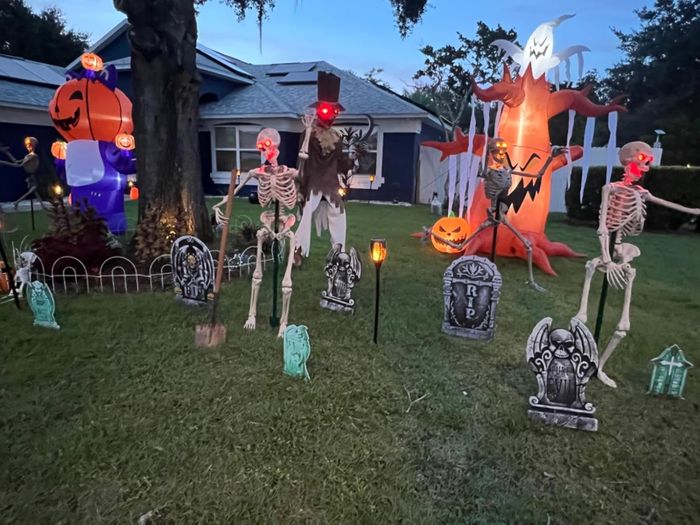A Halloween yard setup showcasing skeletons, tombstones, and large inflatables, including a tree with an owl and a sinister face