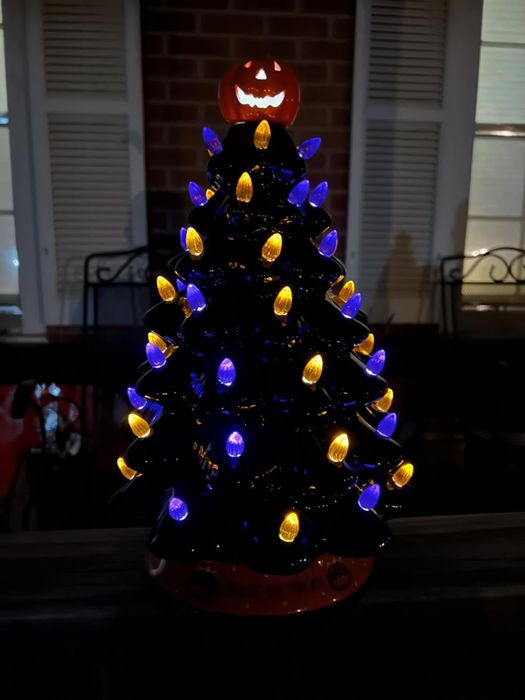 A petite black ornamental tree with glowing orange and purple lights, crowned by a lit jack-o'-lantern, elegantly placed on a table near a window