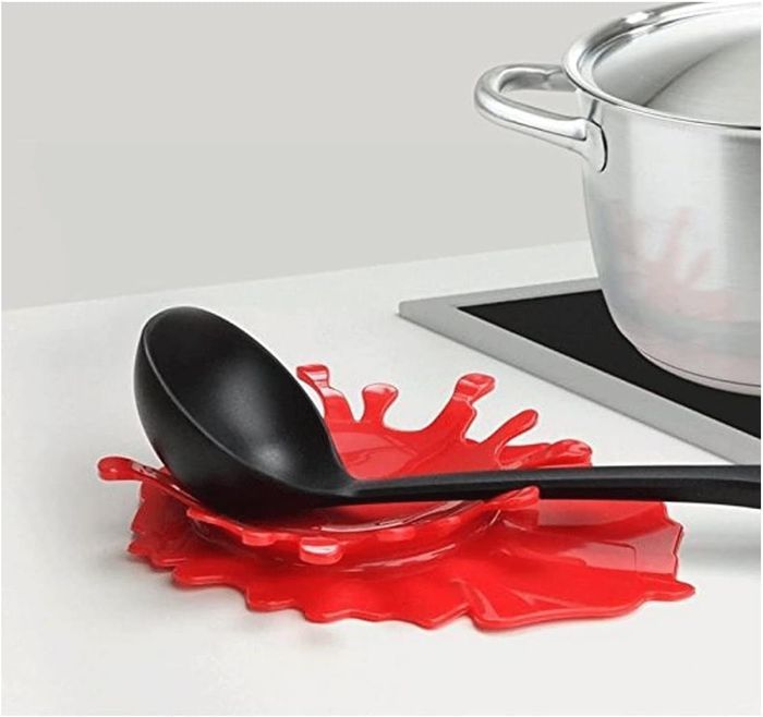 A stainless steel pot on a stove with a black ladle resting on a red, splash-shaped silicone mat