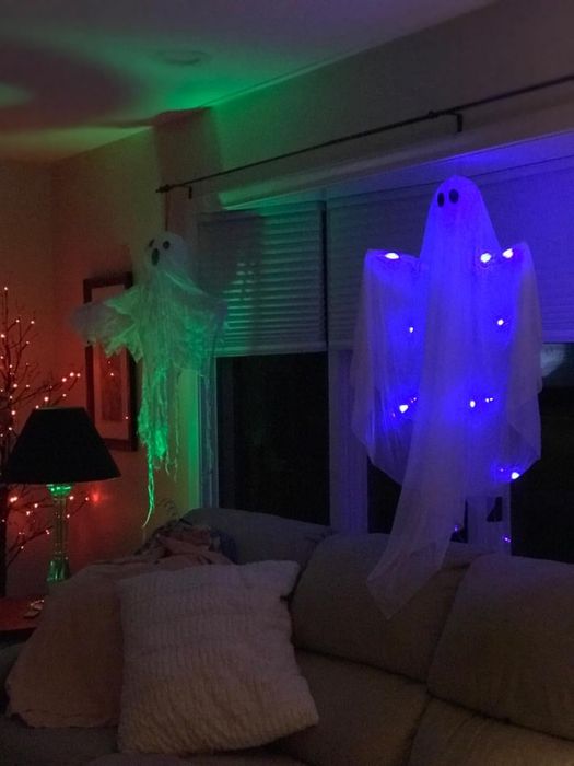 A Halloween-themed room with glowing ghost decorations hanging near windows above a couch, illuminated by purple and green lights