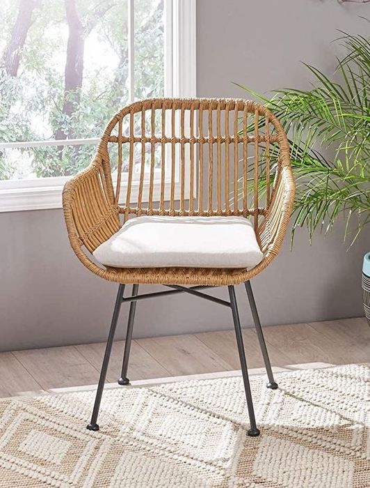 A chic rattan chair with tan woven detailing, complete with a supportive back, armrests, and a cushy tan or white seat, all perched atop sleek black metal legs.