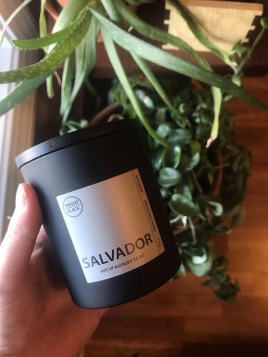 A hand gently holds a candle in a sleek black jar with a label reading 'Salvador'.