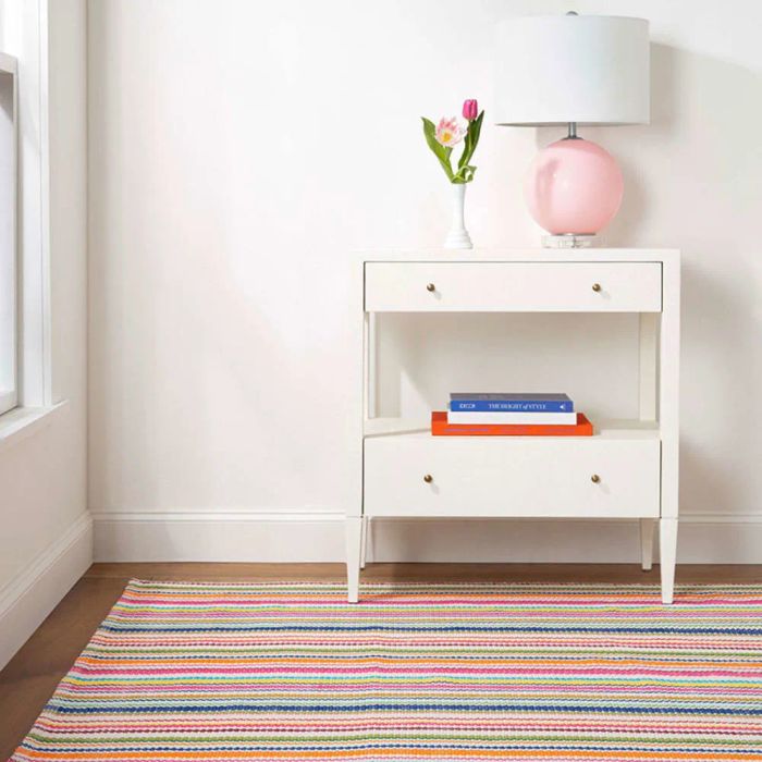 A sleek modern nightstand sits beside a bed, with a pink lamp, a vase of flowers, and two books stacked neatly on one shelf. A striped rug adds a vibrant touch to the floor beneath it.