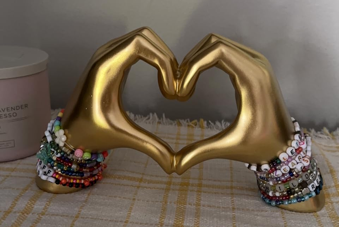 A gold hand sculpture forming a heart, adorned with vibrant beaded bracelets, displayed on a plaid cloth backdrop