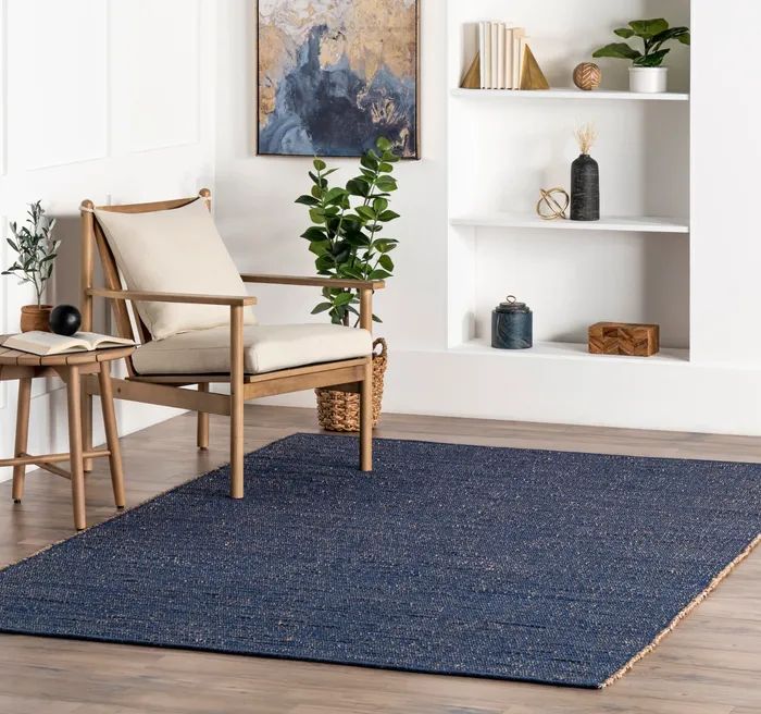 Living room with natural wood chair, small side table, lush plant, and large woven rug on wooden floor; books and decorative items on built-in shelves