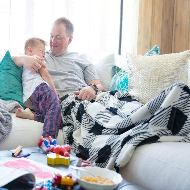 A parent relaxing on the sofa with their legs and feet snuggled under a cozy blanket, while their child curls up beside them