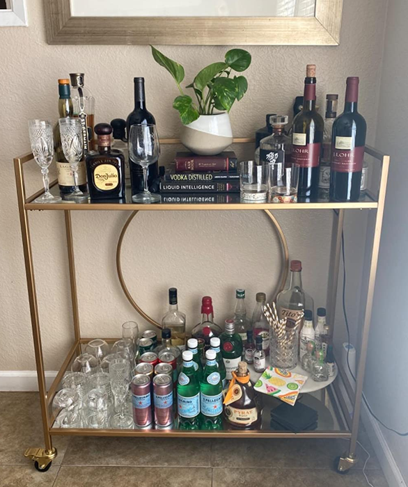 A photo of the gold bar cart filled with bottles and bar accessories, placed against a wall.