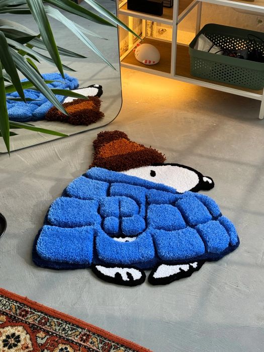 A fun rug shaped like a cartoon character, wearing a puffy blue jacket and brown hat, sits on the floor beside a plant and a shelf, adding whimsy and charm to the space.