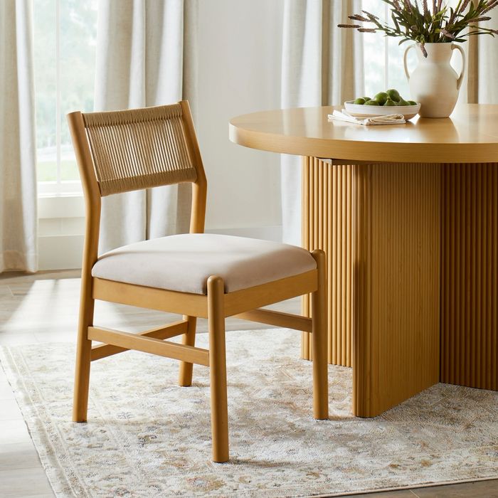 A wooden dining chair featuring a woven backrest and a cushioned seat, positioned next to a rounded wooden table adorned with a vase and a bowl.