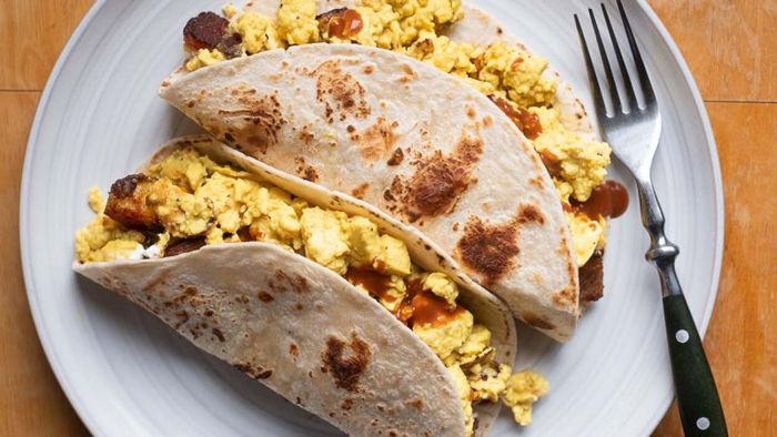 Aerial view of two breakfast tacos nestled in flour tortillas, served on a white plate.