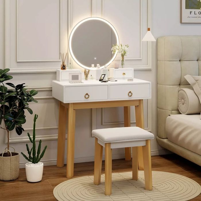 Contemporary vanity table featuring a lighted mirror, wooden legs, white drawers, and a matching stool, set in a chic bedroom adorned with plants and decorative accents