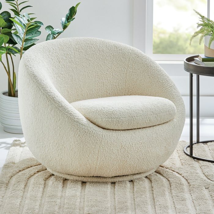 A contemporary armchair with a plush, rounded design, placed on a textured rug with potted plants and a side table nearby, creating a cozy and stylish atmosphere.