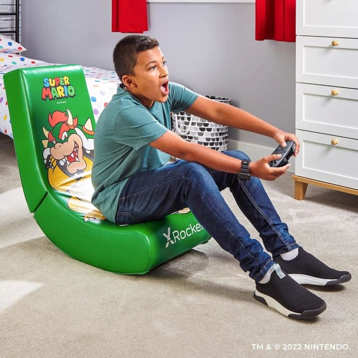 A child immersed in a video game while sitting in a green Super Mario-themed gaming chair, fully focused on the TV. The room features a bed and dresser in the background.