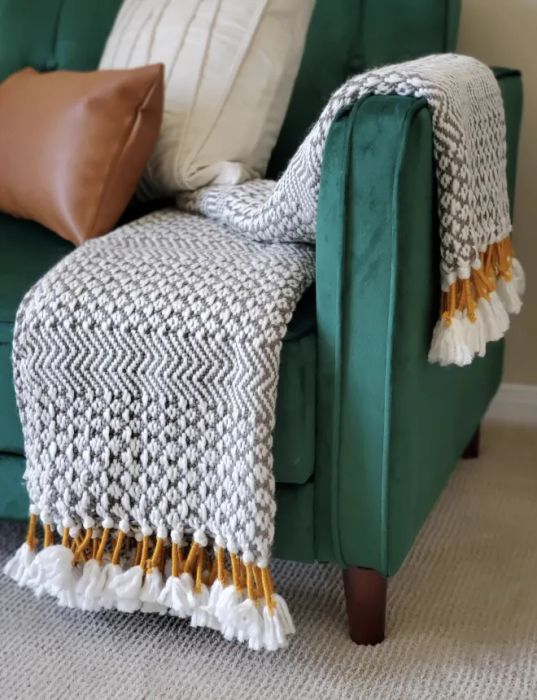 A customer review photo showcasing the woven blanket in grey and white with mustard yellow accents, adding a vibrant touch to any living space.