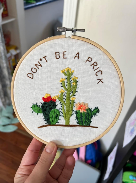 A hand holding an embroidery hoop featuring a design of three cacti with text.