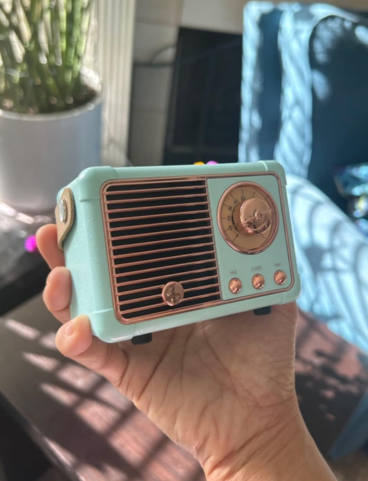 A person holding a small, retro-inspired Bluetooth speaker designed like a vintage radio, complete with a front grill, tuning dial, and three control buttons.