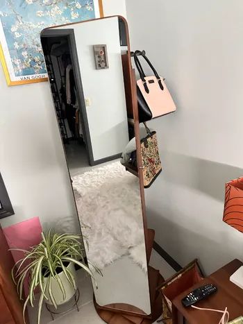 A full-length mirror reflecting a handbag hanging from its corner, set in a room filled with various items
