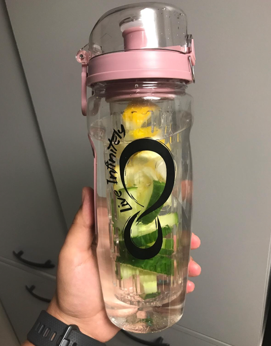 A hand holds a transparent water bottle with a pink lid, infused with slices of cucumber and lemon. The bottle features a black logo on its side.