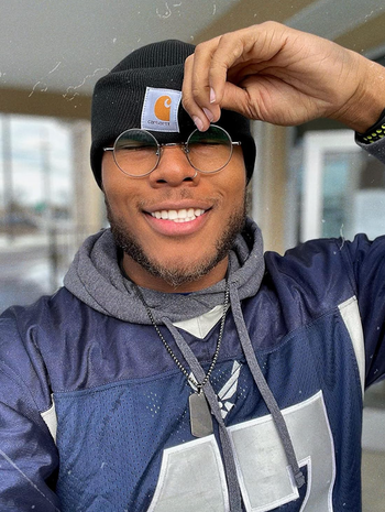 A person grinning while wearing a beanie, glasses, and a casual sports jersey adorned with necklaces, posing outdoors.