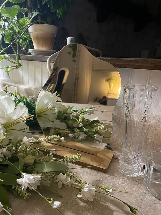 A uniquely shaped mirror resting on a wooden stand, reflecting a delicate arrangement of flowers.