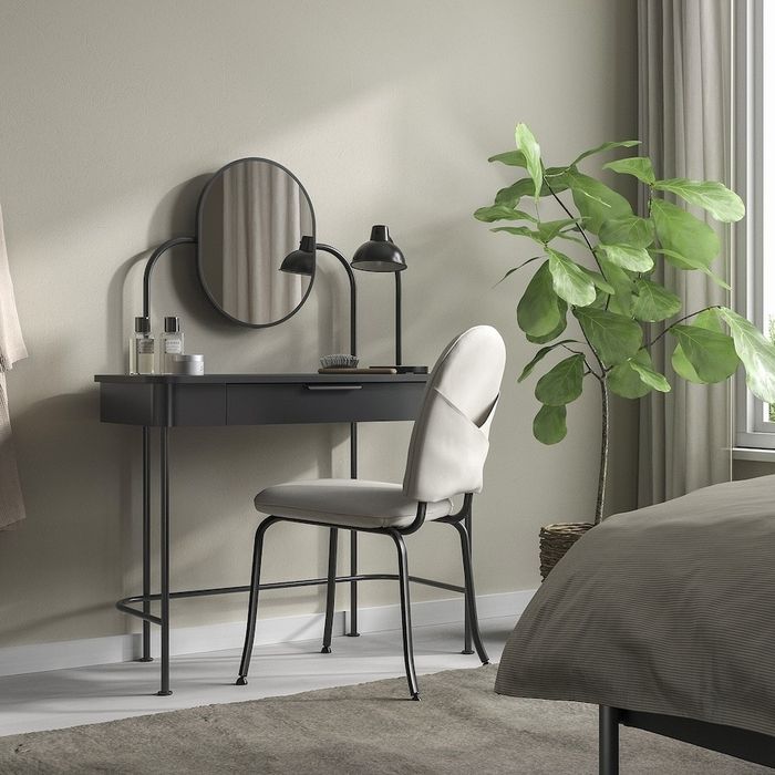 Vanity table featuring a round mirror, black chair, desk lamp, and a plant placed near a window in a cozy bedroom setting