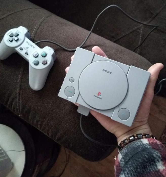 A hand gripping the compact PlayStation console, complete with its games and a standard controller.