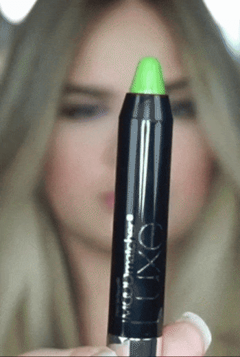 A close-up of a person holding a green-tipped makeup pencil in front of a softly blurred background.