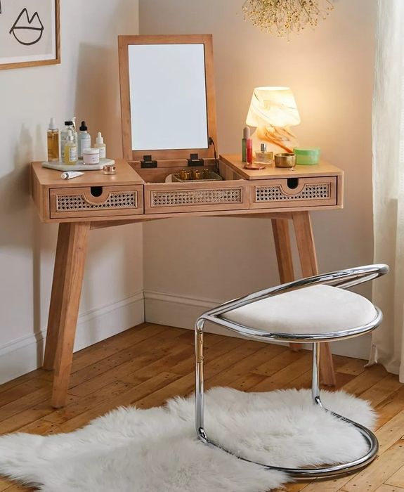 A wooden vanity featuring a mirror, an assortment of beauty products, and a lamp, complemented by a contemporary chrome and fur chair on a wooden floor