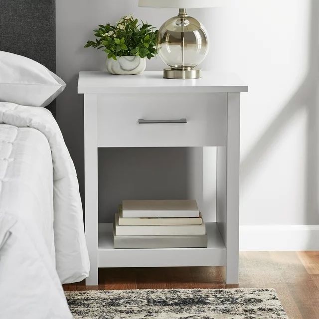 A sleek white nightstand with a drawer, neatly holding books underneath, a gold lamp on top, positioned beside a cozy bed.