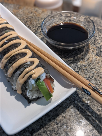 A sushi roll with avocado and carrot, elegantly plated on a white dish, topped with sauce, and accompanied by chopsticks and a side of soy sauce.
