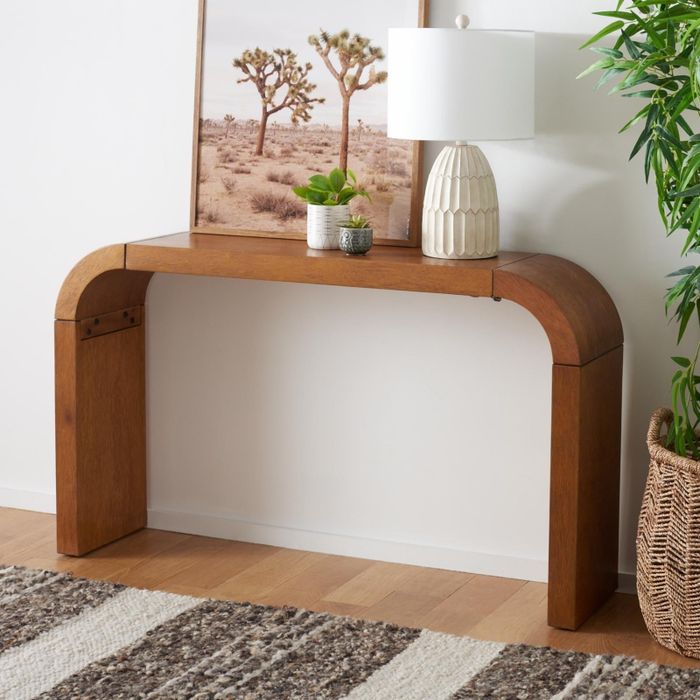 A wooden console table with smooth, rounded edges, decorated with a desert landscape photo, a white lamp, a small plant, and a wicker basket placed on the side.
