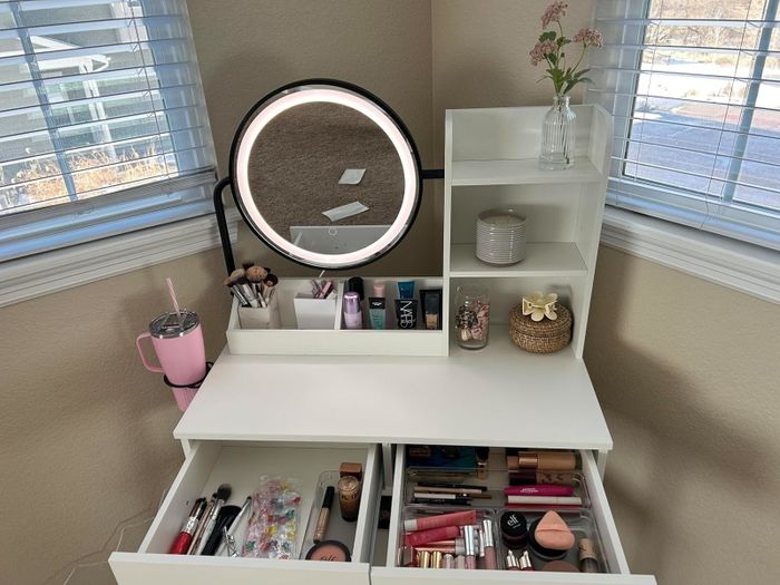 Vanity desk featuring a round mirror, open drawers displaying makeup items, a pink tumbler, and a floral vase