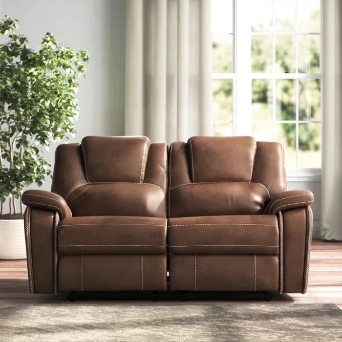 A modern brown leather reclining loveseat is positioned in a sunlit room with expansive windows, complemented by a nearby potted plant.