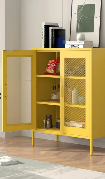Yellow cabinet with glass doors showcasing books, snacks, and decorative items. A small yellow table nearby holds bottles and glasses, with a green potted plant beside the cabinet