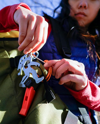 A person attaching a multitool to a backpack, featuring tools like a wrench, ruler, and hex key, perfect for outdoor adventures