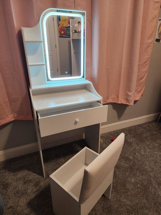 White vanity set with a lighted mirror, storage shelves, an open drawer, and a cushioned stool placed on a carpeted floor, complemented by pink curtains in the background