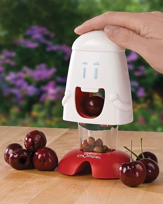 A hand pressing down on a white smiley face tool designed to collect cherry pits, with the clear base showing the collected pits.