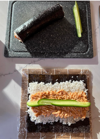 Sushi prep in progress with a bamboo mat, rice, seaweed, spicy salmon, and cucumber slices, along with a completed roll resting on a black cutting board.