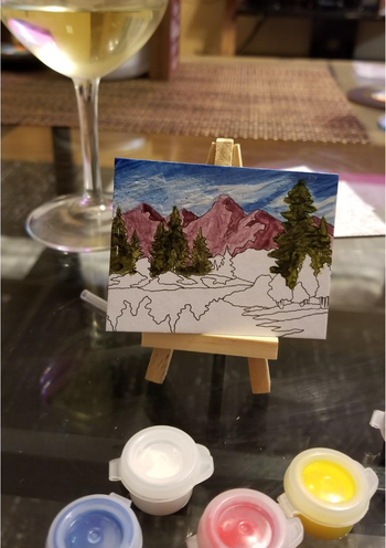 A small landscape painting displayed on an easel, with paint containers and a glass of wine nearby on a table.