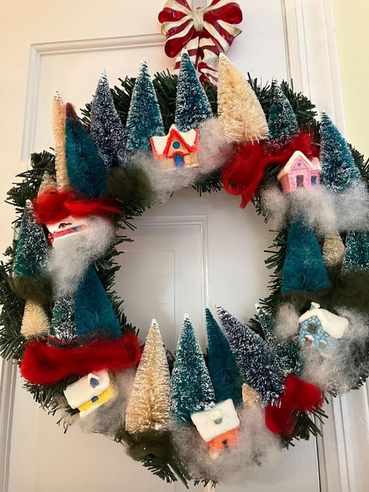 a close-up of a wreath adorned with bottle brush trees and tiny village houses, hanging on a reviewer's front door