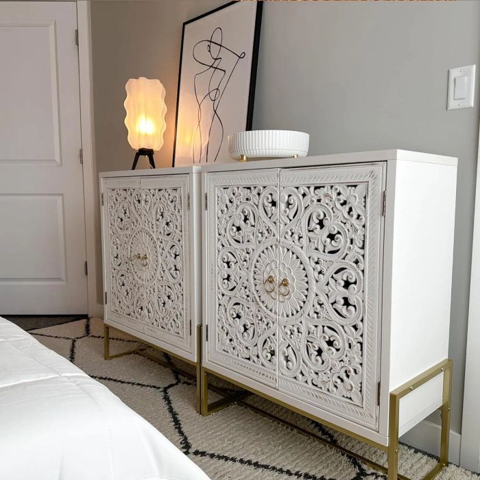ornate white cabinets with detailed designs, topped with a lit lamp, a round bowl, and abstract artwork, positioned next to a white door and a bed