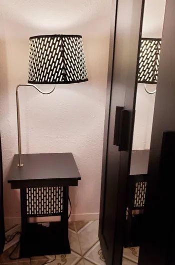 a modern floor lamp with a geometric-patterned shade, placed next to a small black side table, reflected in a mirrored closet door. The scene gives off a vibe of home decor shopping.