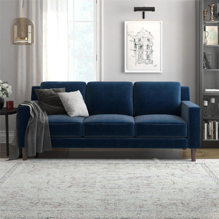 A chic modern living room featuring a deep blue velvet sofa, cozy throw pillows, and a soft gray blanket. Stylish wall art and a neatly arranged bookshelf complete the aesthetic.