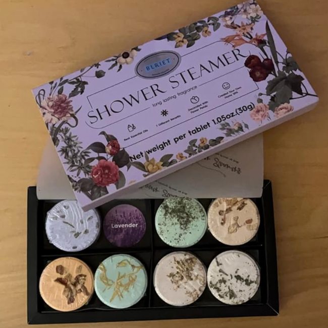 A box of seven shower steamers in various scents, displayed on a wooden surface. The label on the box includes