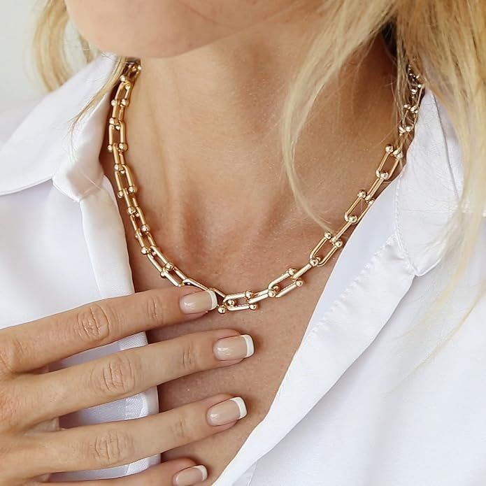 A close-up shot of someone wearing a white shirt and a sleek gold chain necklace. Their fingers, adorned with a French manicure, gently rest on the necklace.