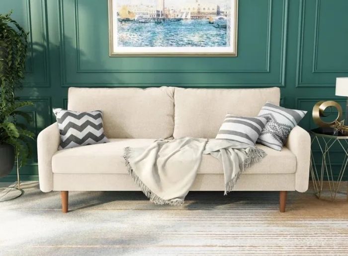 A sleek beige sofa, adorned with three decorative pillows and a cozy throw blanket, sets the tone in a contemporary living room, complete with a painting and a lush plant.