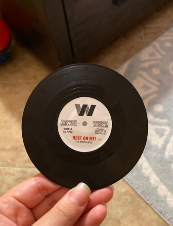 A black mini vinyl record coaster held in a reviewer’s hand, featuring the text 