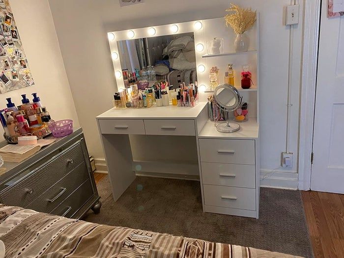 Makeup vanity with a lighted mirror, multiple drawers, and shelves showcasing neatly arranged cosmetics, perfumes, and decorative accents